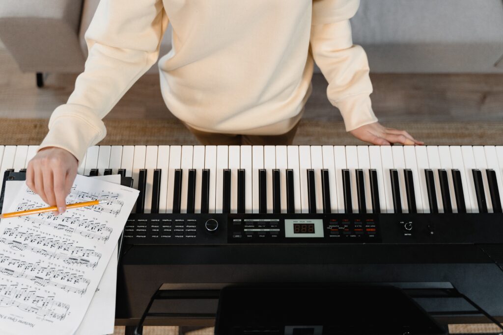 A birds-eye-view of someone sitting at a digital keyboard. There is sheet music resting on the keyboard and the person is holding a pencil over it.